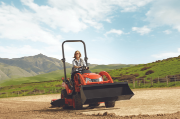 Žena řídí červený traktor Kioti na obdělaném poli se zelenými kopci v pozadí.